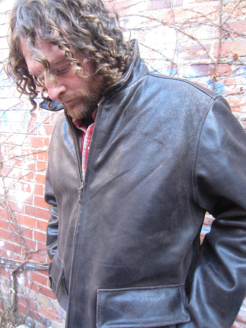 May include: A dark brown leather jacket with a zipper closure and two front pockets. The jacket has a collar and is worn over a red and white plaid shirt. The person wearing the jacket is standing in front of a brick wall.