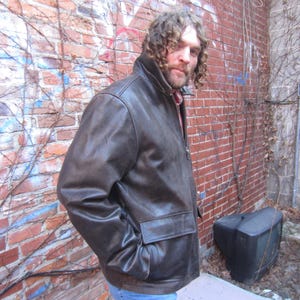 May include: A brown leather jacket with a zip-up front and two front pockets. The jacket has a collar and is worn over a red and white plaid shirt. The person is wearing blue jeans.
