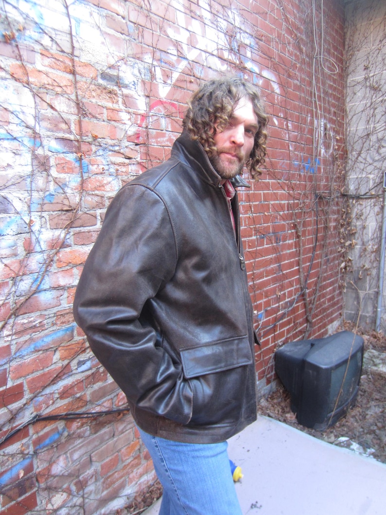 May include: A brown leather jacket with a zip-up front and two front pockets. The jacket has a collar and is worn over a red and white plaid shirt. The person is wearing blue jeans.