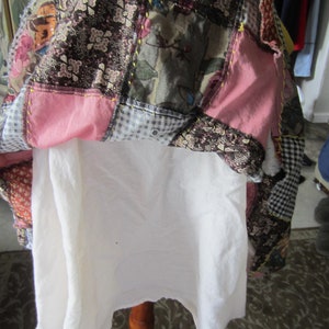 Op de afbeelding: Een patchworkdeken met verschillende stoffen en kleuren, waaronder roze, bruin en wit. De deken is dubbelgevouwen en gedrapeerd over een witte stoffen standaard.