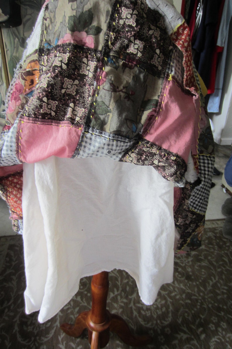 Op de afbeelding: Een patchworkdeken met verschillende stoffen en kleuren, waaronder roze, bruin en wit. De deken is dubbelgevouwen en gedrapeerd over een witte stoffen standaard.