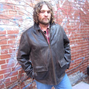 May include: A brown leather jacket with a zip-up front and two front pockets. The jacket is worn over a red and black plaid shirt and blue jeans. The person is standing in front of a brick wall with some graffiti.