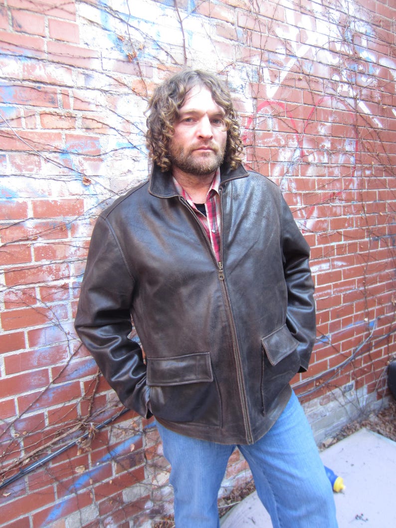 May include: A brown leather jacket with a zip-up front and two front pockets. The jacket is worn over a red and black plaid shirt and blue jeans. The person is standing in front of a brick wall with some graffiti.