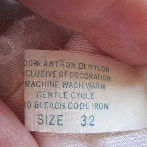 Pode incluir: Etiqueta de cuidado de tecido branca com texto azul. A etiqueta diz "100% Antron III Nylon, Exclusive of Decoration, Machine Wash Warm, Gentle Cycle, No Bleach Cool Iron, Size 32."