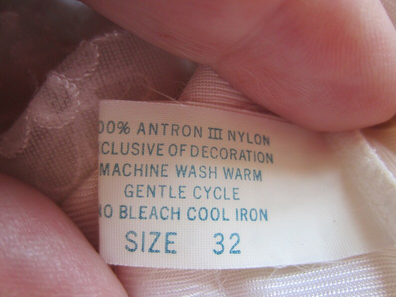 Pode incluir: Etiqueta de cuidado de tecido branca com texto azul. A etiqueta diz "100% Antron III Nylon, Exclusive of Decoration, Machine Wash Warm, Gentle Cycle, No Bleach Cool Iron, Size 32."