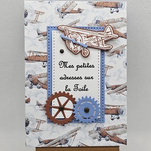 Peut inclure: Une carte décorative avec un motif d'avion vintage. La carte a une bordure bleue et un fond avec des illustrations d'avions répétées. Le texte "Mes petites adresses sur la Toile" est affiché sur la carte. Un petit chevalet en bois soutient la carte.