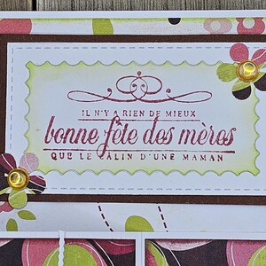 May include: A handmade Mother's Day card with a floral design. The card features a rectangular frame with the text "bonne f&ecirc;te des m&egrave;res" in burgundy. Decorative flowers with gold accents are placed around the frame.