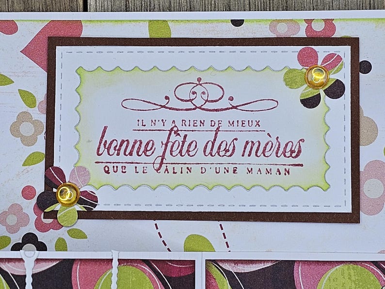 May include: A handmade Mother's Day card with a floral design. The card features a rectangular frame with the text "bonne f&ecirc;te des m&egrave;res" in burgundy. Decorative flowers with gold accents are placed around the frame.