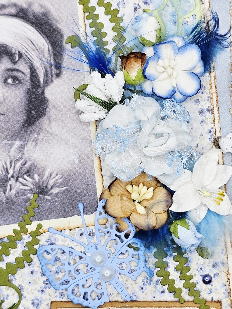 May include: A vintage-style scrapbook page featuring a black and white portrait of a woman. The page is adorned with blue and white paper flowers, blue feathers, and a light blue butterfly cutout with pearl accents. Green decorative edges frame the design.