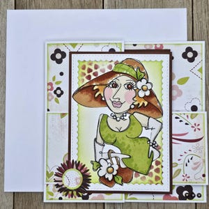 May include: A handmade greeting card featuring a whimsical illustration of a woman in a hat and dress. The card has a layered design with floral and patterned paper accents. The woman is wearing a green dress and a brown hat.