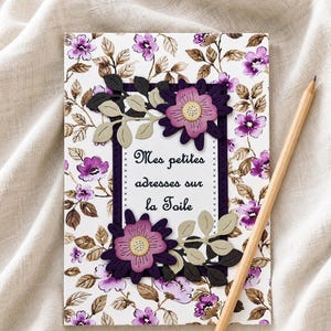 Peut inclure: Un carnet à motifs floraux avec le texte "Mes petites adresses sur la Toile" est présenté avec un crayon en bois. Le carnet présente un motif floral violet et blanc avec des fleurs et des feuilles décoratives violettes. Le crayon est marron clair avec une bande dorée.