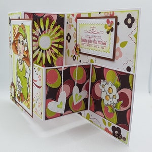 May include: A handmade Mother's Day card with a unique tri-fold design. The card features a cartoon illustration of a woman, floral patterns, and the French text "bonne f&ecirc;te des m&egrave;res". The color palette includes shades of green, brown, pink, and white.
