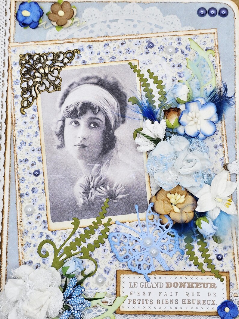 May include: A vintage-style scrapbook page with a black and white portrait of a woman. The page features blue floral patterned paper, lace, and decorative elements like flowers, butterflies, and a quote in French.