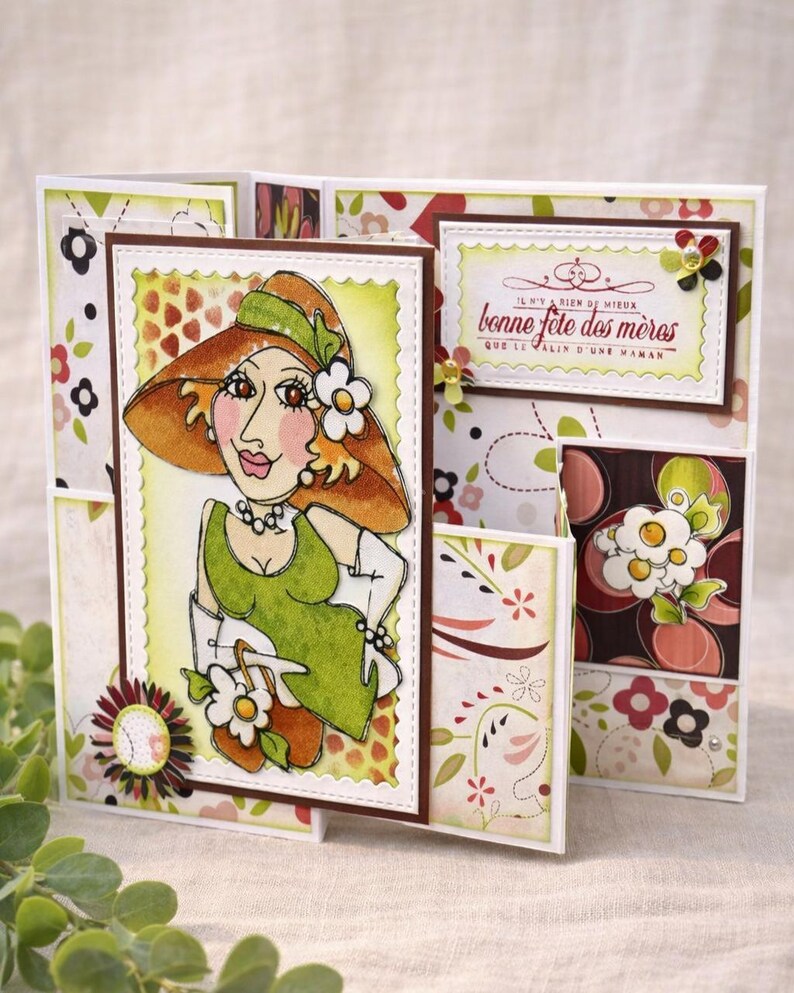 May include: A handmade greeting card featuring a cartoon illustration of a woman in a hat and dress. The card has a floral pattern and includes the text "Bonne F&ecirc;te des M&egrave;res". The card is multi-panel and decorated with embellishments.