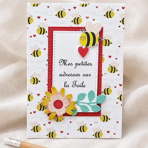 Peut inclure: Une carte de vœux sur le thème des abeilles. La carte présente un fond blanc avec des abeilles jaunes et noires répétées et des cœurs rouges. La carte a une bordure rouge avec le texte français "Mes petites adresses sur la Toile". Un crayon repose en bas.