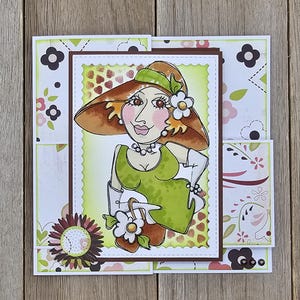 May include: A handmade greeting card featuring a cartoon illustration of a woman in a green dress and a brown hat. The card has a layered design with floral and patterned paper accents in shades of green, brown, and pink.