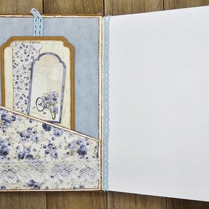 May include: Open journal with a light blue and floral design. The left side features a pocket with a floral pattern and lace trim. Inside, there are decorative tags with a bicycle illustration. The right side is blank.