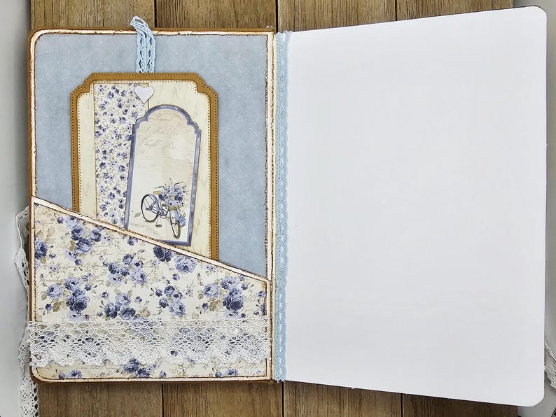 May include: Open journal with a light blue and floral design. The left side features a pocket with a floral pattern and lace trim. Inside, there are decorative tags with a bicycle illustration. The right side is blank.