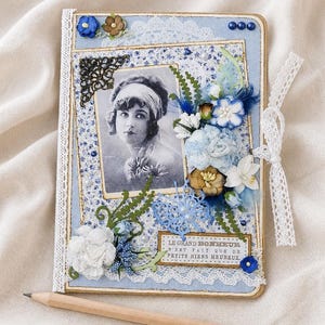 May include: A handmade scrapbook with a vintage photograph of a woman, adorned with blue and white floral embellishments, lace trim, and a decorative butterfly. The text "LE GRAND BONHEUR N'EST FAIT QUE DE PETITS RIENS HEUREUX" is visible. A pencil rests below.