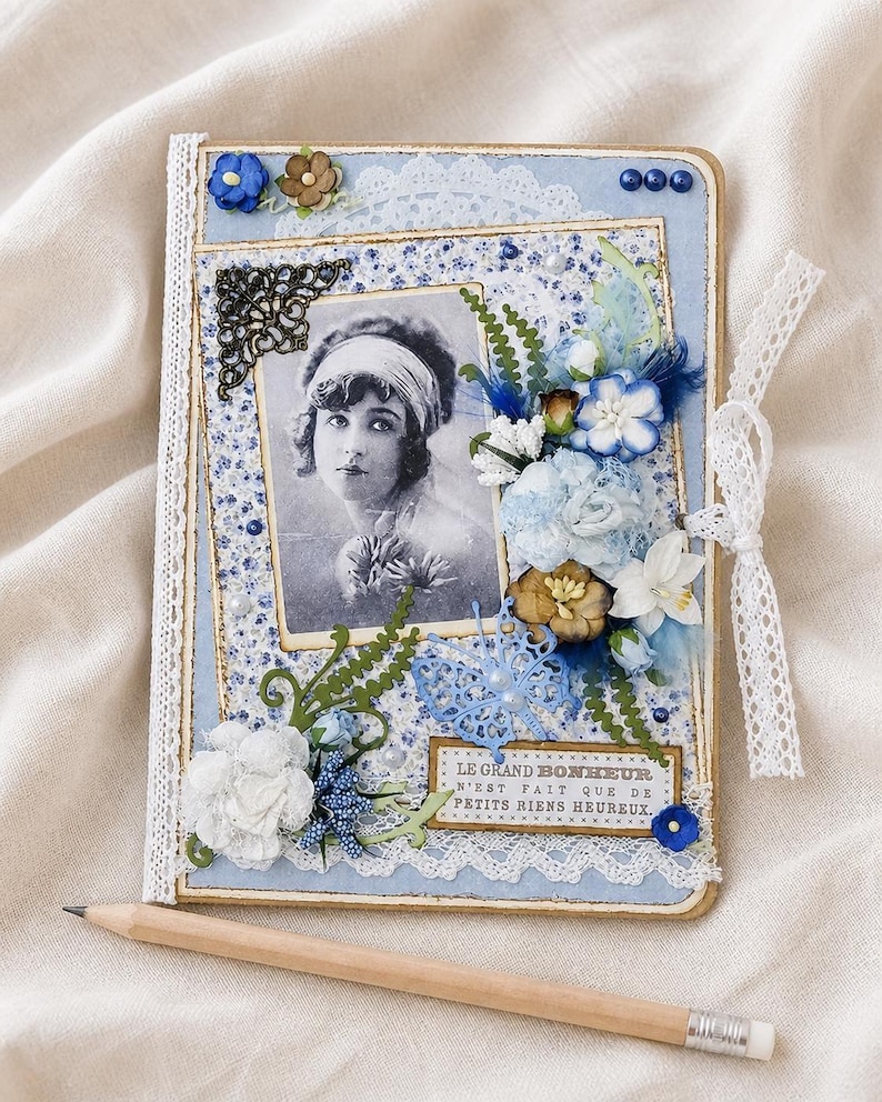 May include: A handmade scrapbook with a vintage photograph of a woman, adorned with blue and white floral embellishments, lace trim, and a decorative butterfly. The text "LE GRAND BONHEUR N'EST FAIT QUE DE PETITS RIENS HEUREUX" is visible. A pencil rests below.