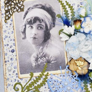 May include: A vintage-style scrapbook page featuring a black and white portrait of a woman. The page is decorated with blue and white floral patterned paper, paper flowers, a blue butterfly, and green paper cutouts. A decorative bronze corner piece adds detail.