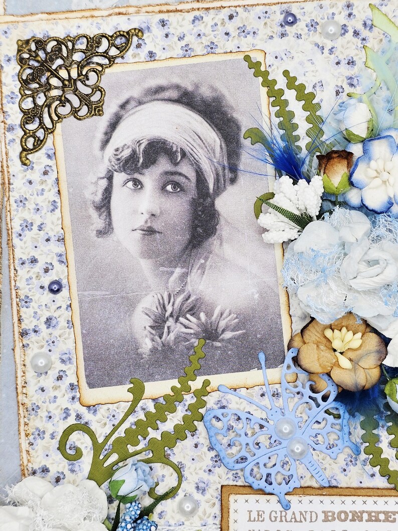 May include: A vintage-style scrapbook page featuring a black and white portrait of a woman. The page is decorated with blue and white floral patterned paper, paper flowers, a blue butterfly, and green paper cutouts. A decorative bronze corner piece adds detail.