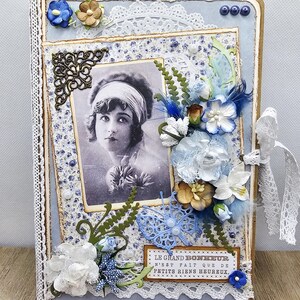 May include: A vintage-style scrapbook with a portrait of a woman. The cover is light blue with floral and lace embellishments, including paper flowers, a butterfly, and the text "LE GRAND BONHEUR N'EST FAIT QUE DE PETITS RIENS HEUREUX."