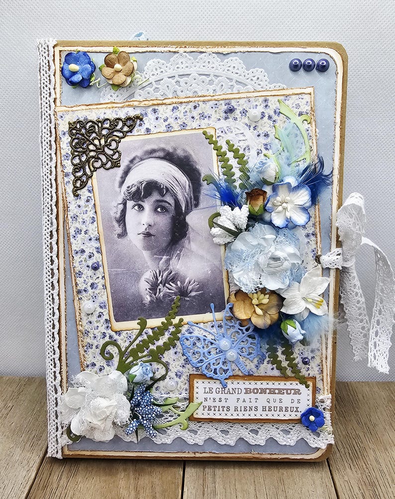 May include: A vintage-style scrapbook with a portrait of a woman. The cover is light blue with floral and lace embellishments, including paper flowers, a butterfly, and the text "LE GRAND BONHEUR N'EST FAIT QUE DE PETITS RIENS HEUREUX."