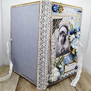 May include: A handmade scrapbook album with a vintage aesthetic. The album features a light blue cover with a vertical ribbed texture, adorned with white lace trim and a decorative photo frame. The frame contains a black and white portrait, embellished with blue and white floral accents.