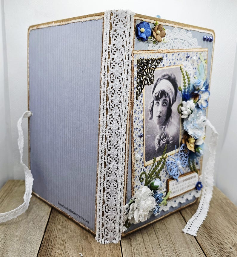 May include: A handmade scrapbook album with a vintage aesthetic. The album features a light blue cover with a vertical ribbed texture, adorned with white lace trim and a decorative photo frame. The frame contains a black and white portrait, embellished with blue and white floral accents.