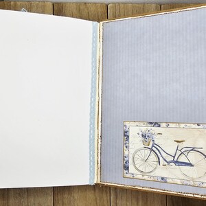 May include: Open journal with blank white pages and a light blue patterned page. A small framed image of a blue bicycle with a basket of flowers is on the right page. The journal has a light blue lace trim.