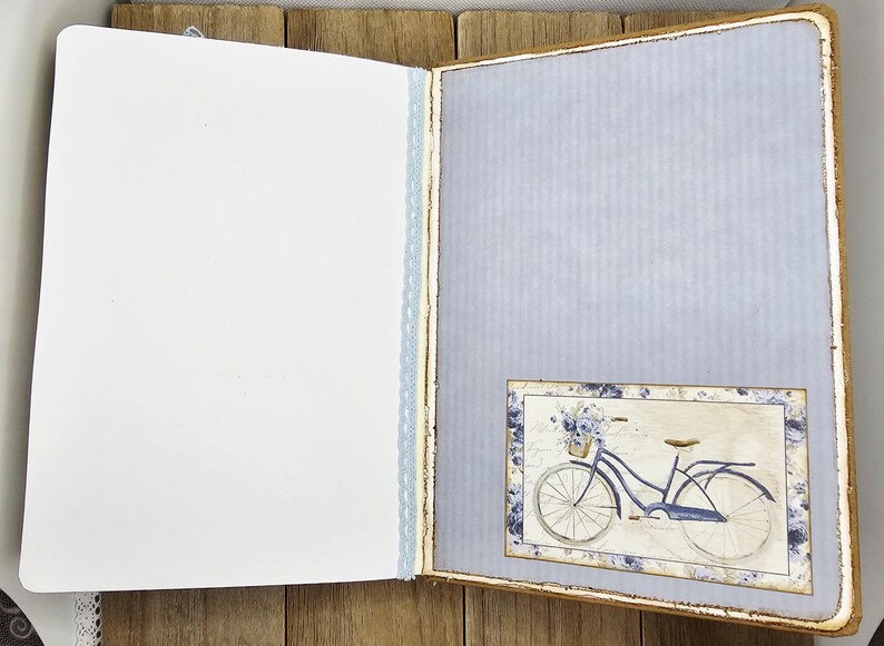 May include: Open journal with blank white pages and a light blue patterned page. A small framed image of a blue bicycle with a basket of flowers is on the right page. The journal has a light blue lace trim.