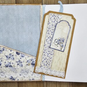 May include: Open journal with a light blue interior and a pocket with a floral pattern in blue and white. A decorative tag with a bicycle illustration and lace trim is attached. The journal is open on a wooden surface.