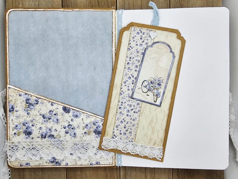 May include: Open journal with a light blue interior and a pocket with a floral pattern in blue and white. A decorative tag with a bicycle illustration and lace trim is attached. The journal is open on a wooden surface.