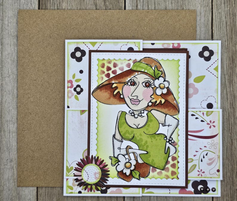 May include: A handmade greeting card featuring a whimsical illustration of a woman in a hat and dress. The card has a brown border and a floral design. The woman is wearing a green dress and holding a white purse with a flower.