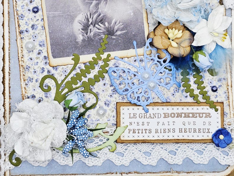 May include: A handmade scrapbook page with a vintage photo, floral embellishments, and a blue butterfly cutout. The page features white lace, blue and white floral patterned paper, and the French text "LE GRAND BONHEUR N'EST FAIT QUE DE PETITS RIENS HEUREUX."