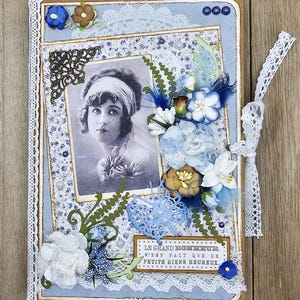 May include: A handmade scrapbook with a vintage aesthetic. The cover features a black and white photo of a woman, surrounded by blue and white floral paper, lace, and decorative embellishments. A ribbon tie secures the book.