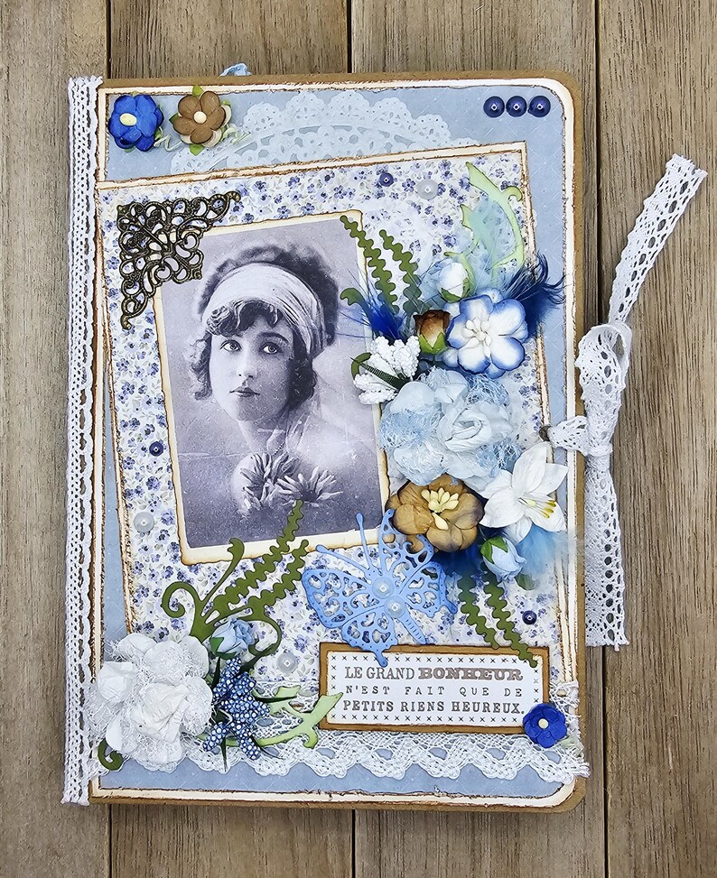 May include: A handmade scrapbook with a vintage aesthetic. The cover features a black and white photo of a woman, surrounded by blue and white floral paper, lace, and decorative embellishments. A ribbon tie secures the book.