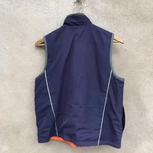 May include: A navy blue vest with white piping and a zipper closure. The vest is hanging on a hanger.