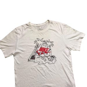 Op de afbeelding: Wit T-shirt met een zwart en rood Nike-logo. De afbeelding toont een paar Nike-schoenen in een hangmat tussen twee palmbomen.
