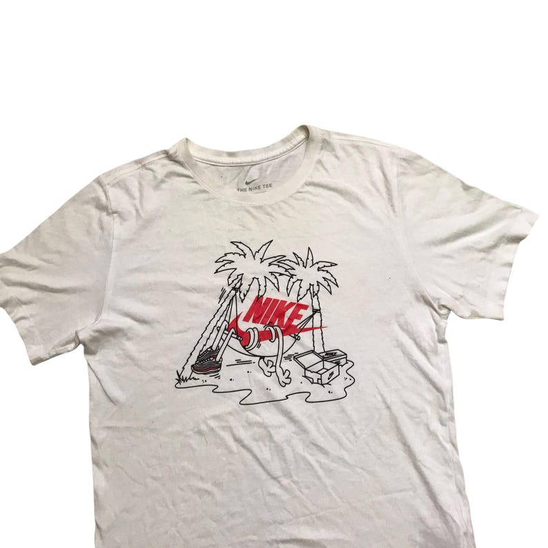 Op de afbeelding: Wit T-shirt met een zwart en rood Nike-logo. De afbeelding toont een paar Nike-schoenen in een hangmat tussen twee palmbomen.