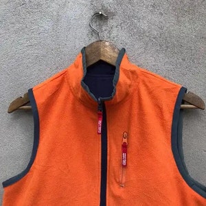May include: Orange fleece vest with a navy blue zipper and trim. The vest has two red zipper pulls.