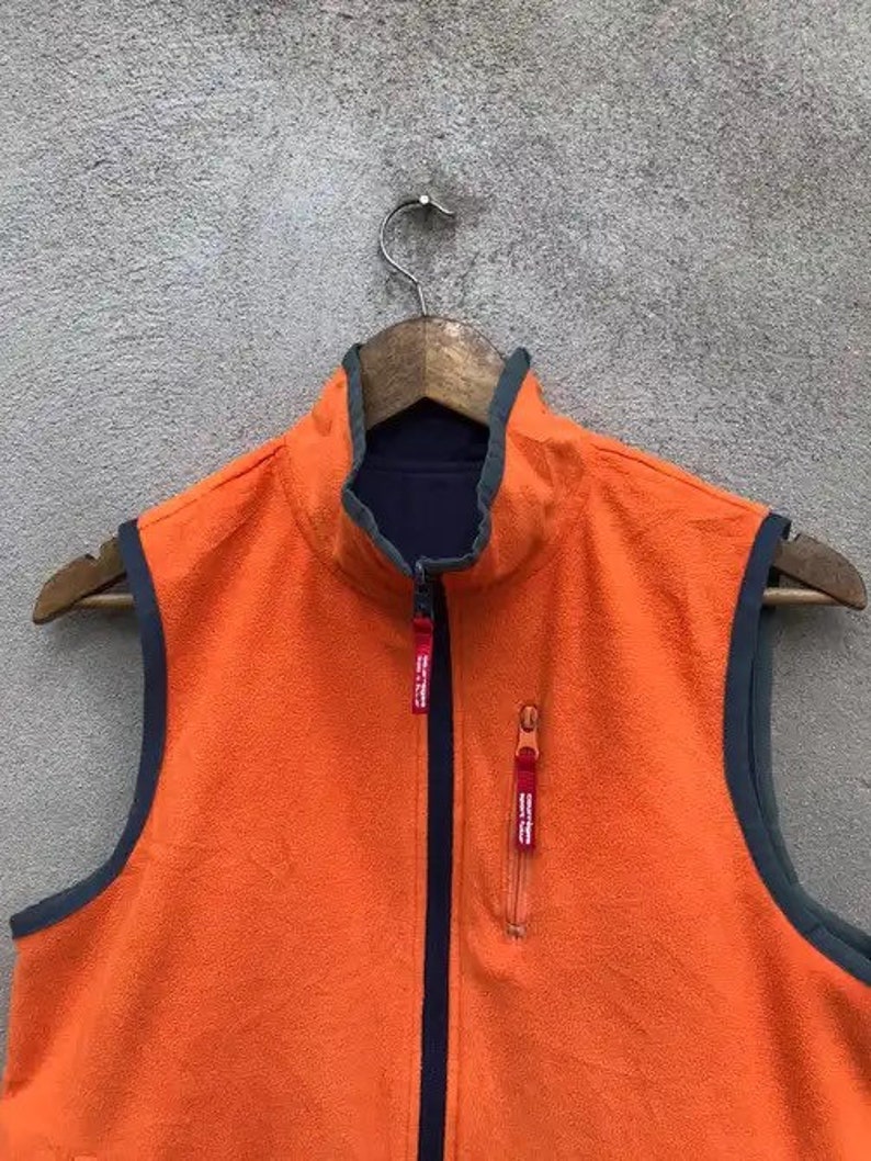 May include: Orange fleece vest with a navy blue zipper and trim. The vest has two red zipper pulls.