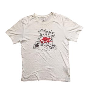 Op de afbeelding: Wit T-shirt met een zwart en rood Nike-logo-ontwerp. Het ontwerp toont een Nike-swoosh met benen en armen die ontspannen in een hangmat tussen twee palmbomen.
