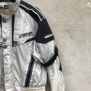 K&ouml;nnte beinhalten: Eine silberne und schwarze Jacke mit einem CYBER-TEX-Logo auf der Vorderseite. Die Jacke hat einen Rei&szlig;verschluss und einen schwarzen Riemen am linken &Auml;rmel.