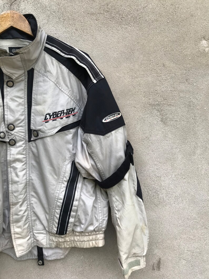 K&ouml;nnte beinhalten: Eine silberne und schwarze Jacke mit einem CYBER-TEX-Logo auf der Vorderseite. Die Jacke hat einen Rei&szlig;verschluss und einen schwarzen Riemen am linken &Auml;rmel.