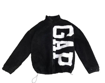 May include: Black fleece jacket featuring the word "GAP" in large white letters. The jacket has a high collar, long sleeves, and a drawstring at the hem. The jacket is made of a soft, fuzzy material, suitable for cooler weather.