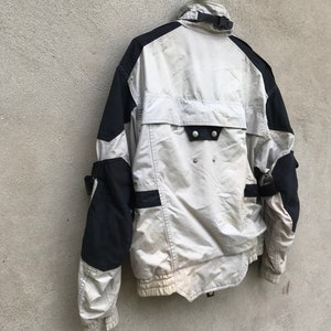 K&ouml;nnte beinhalten: Eine wei&szlig;e und schwarze Jacke mit Rei&szlig;verschluss und einem schwarzen Riemen am Kragen. Die Jacke hat ein schwarz-wei&szlig;es Design an den &Auml;rmeln und eine schwarze Tasche auf der R&uuml;ckseite.
