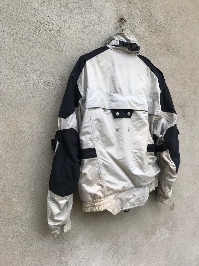 K&ouml;nnte beinhalten: Eine wei&szlig;e und schwarze Jacke mit Rei&szlig;verschluss und einem schwarzen Riemen am Kragen. Die Jacke hat ein schwarz-wei&szlig;es Design an den &Auml;rmeln und eine schwarze Tasche auf der R&uuml;ckseite.