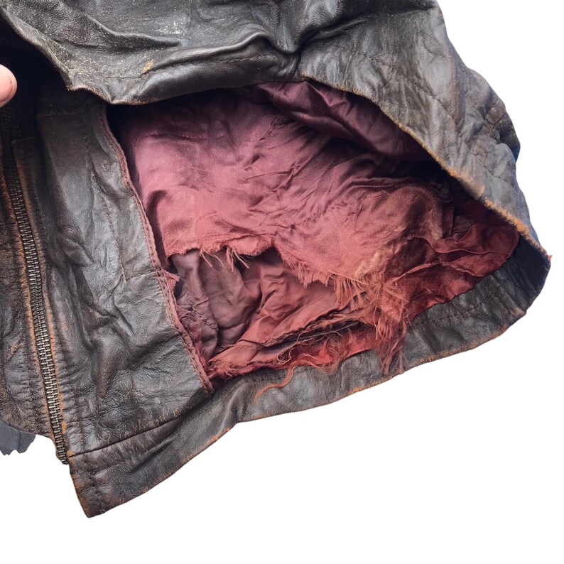 以下が含まれることがあります： 赤い裏地が付いた茶色の革ジャンパー。裏地は破れてほつれています